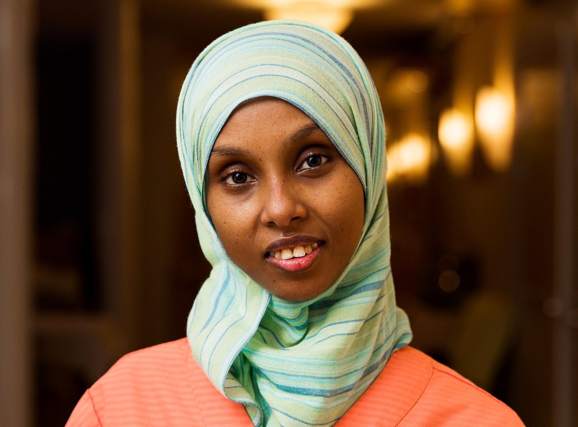 Lähihoitaja Ikran Adan työvaatteissa, vaaleanvihreässä ja siniraidallisessa hijabissa, hymyilee ystävällisesti kameralle. Tausta on lämmin ja epätarkka, mikä luo rauhallisen ja kutsuvan tunnelman.